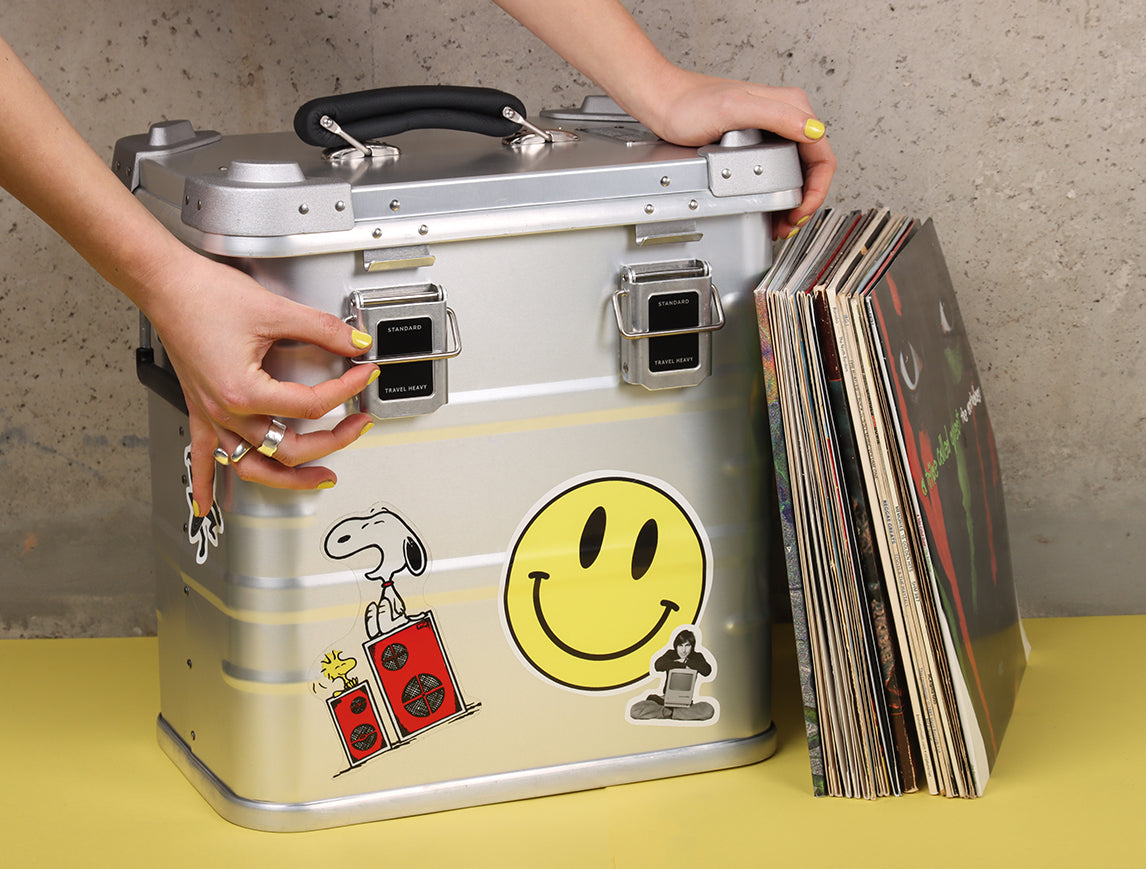 Hands flicking through vinyl records in an open Travel Heavy aluminium record case covered in stickers
